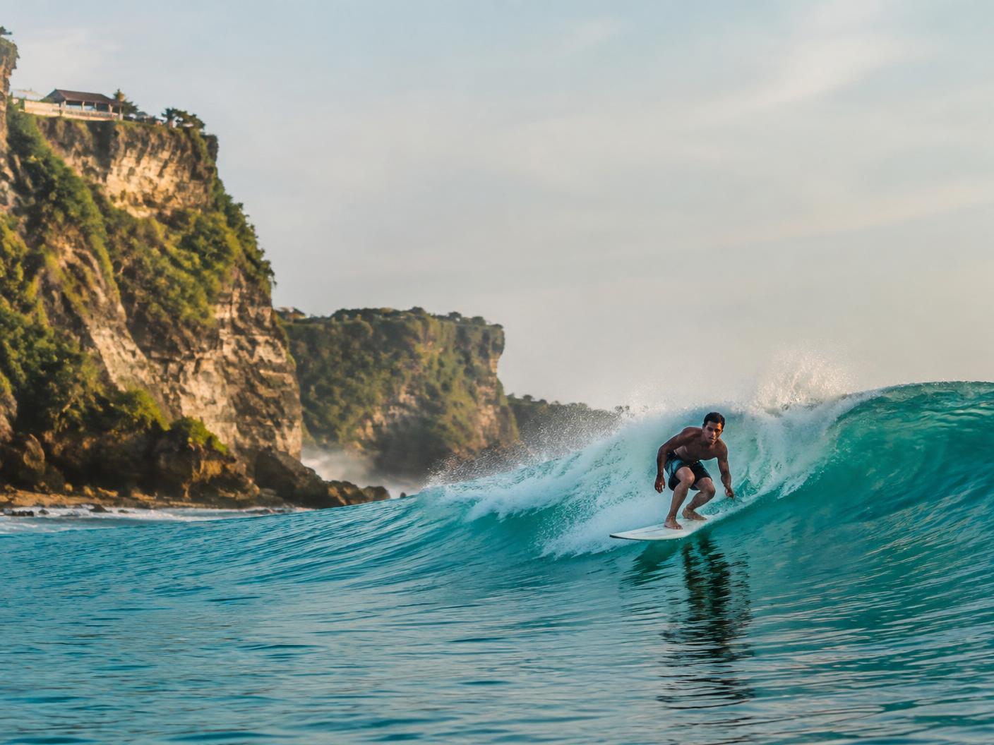 A surfer on a turquoise wave below limestone cliffs